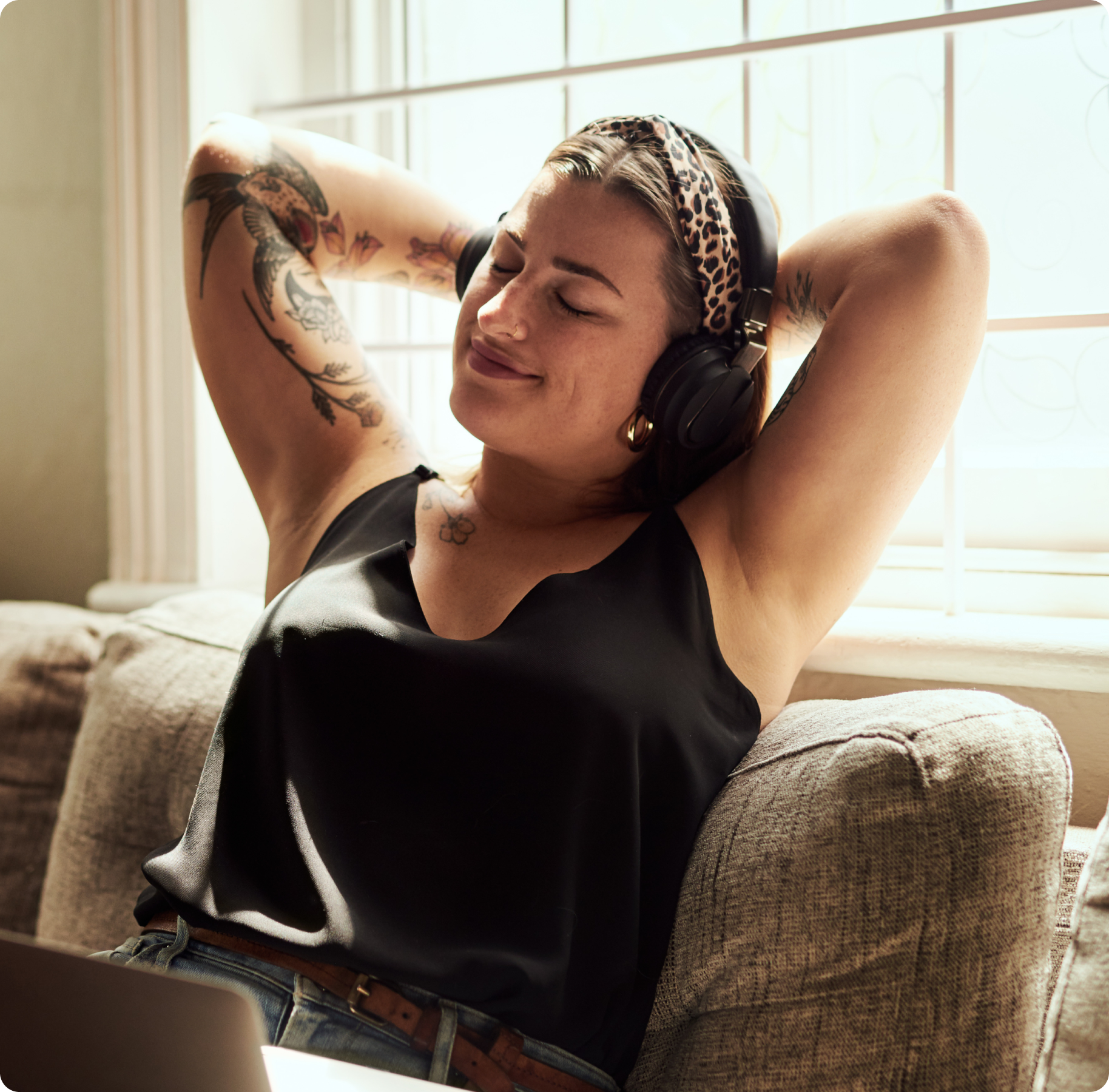 A person relaxes on a couch, wearing headphones and a leopard-print headband, eyes closed, smiling contentedly. Sunlight filters through a window. Tattoos on arms.