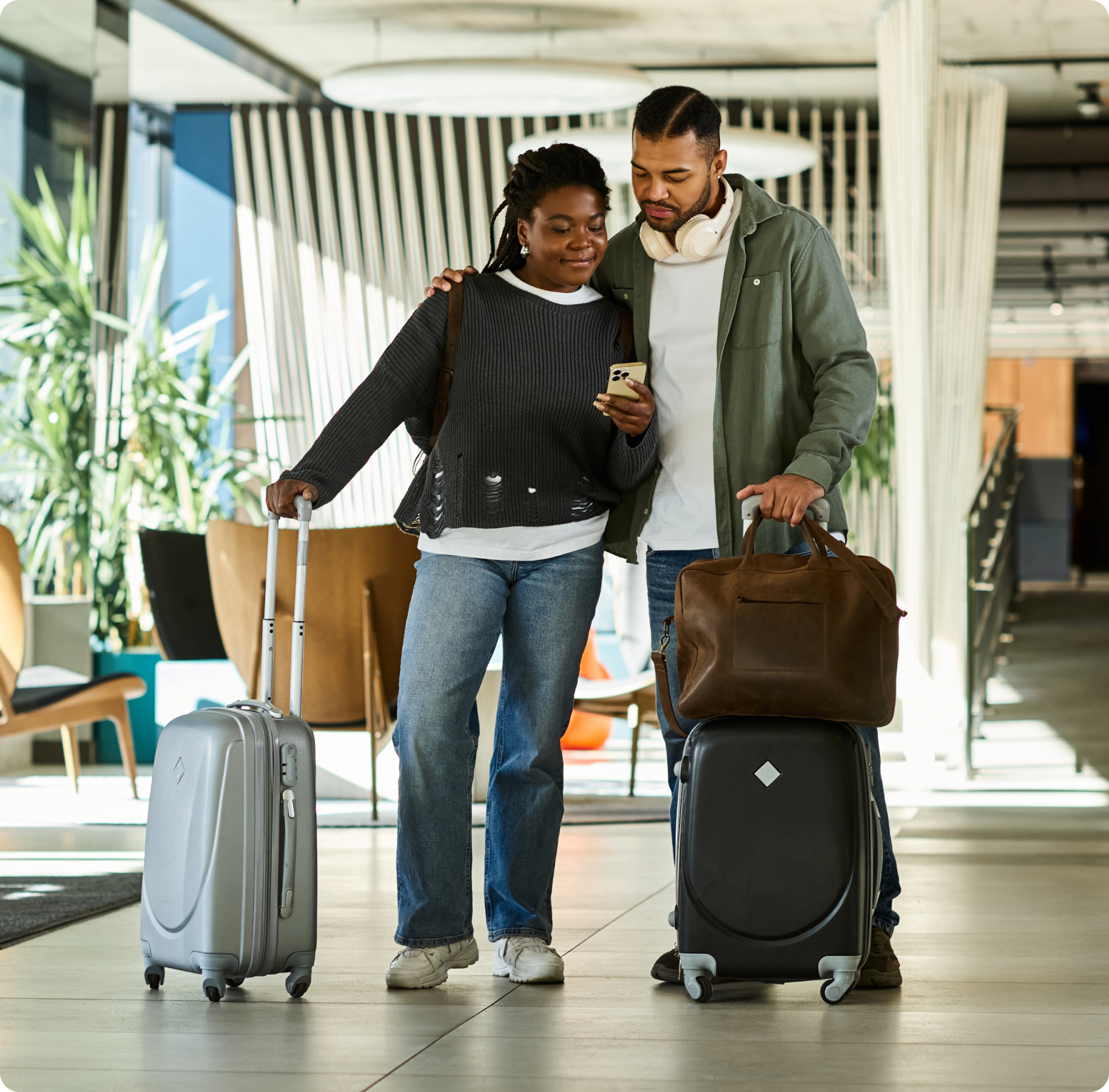 A couple stands in a modern lobby, smiling at a phone. They have luggage, suggesting travel. The scene conveys a relaxed, happy atmosphere.