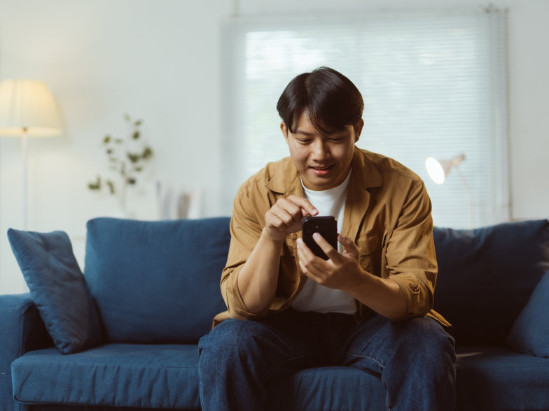 Man in a tan overshirt sitting on a blue couch at home, smiling as he shops on his phone