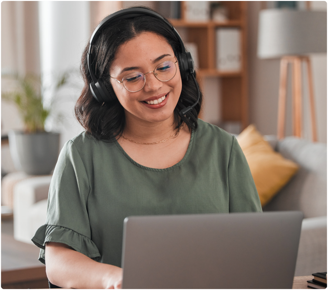 A woman wearing glasses and headphones smiles while using a laptop in a cozy, well-lit living room with plants and a sofa in the background.