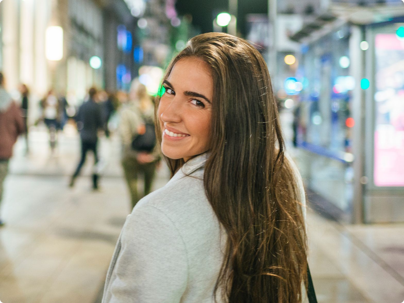 Smiling woman with long brown hair looks back at the camera while walking in a lively, brightly illuminated city street filled with blurred pedestrians.