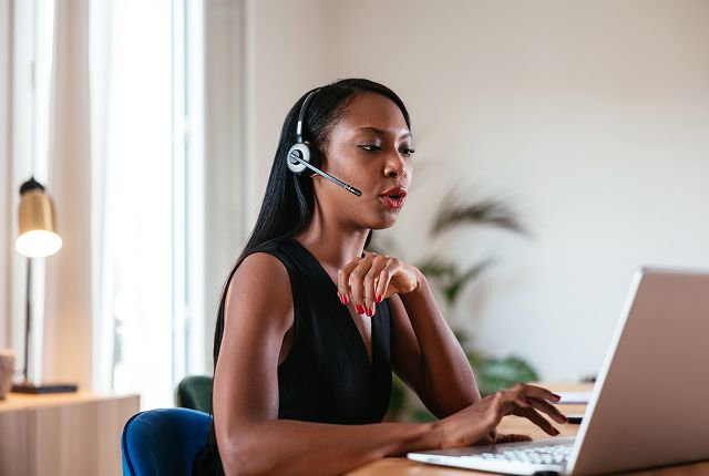 Woman wearing a headset sits at a desk, typing on a laptop in a bright room. She appears focused and engaged, suggesting a professional setting.