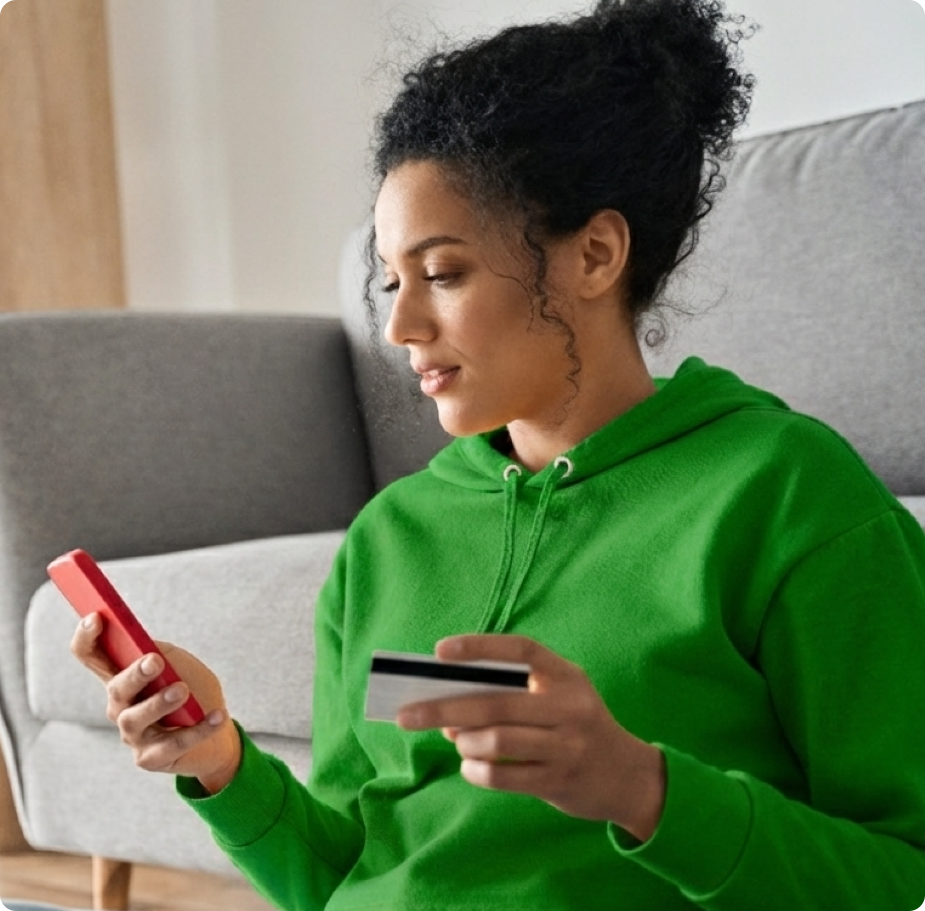 A woman in a green hoodie holds a smartphone and a credit card, sitting on a sofa at home. She appears focused, engaged in online shopping or banking.