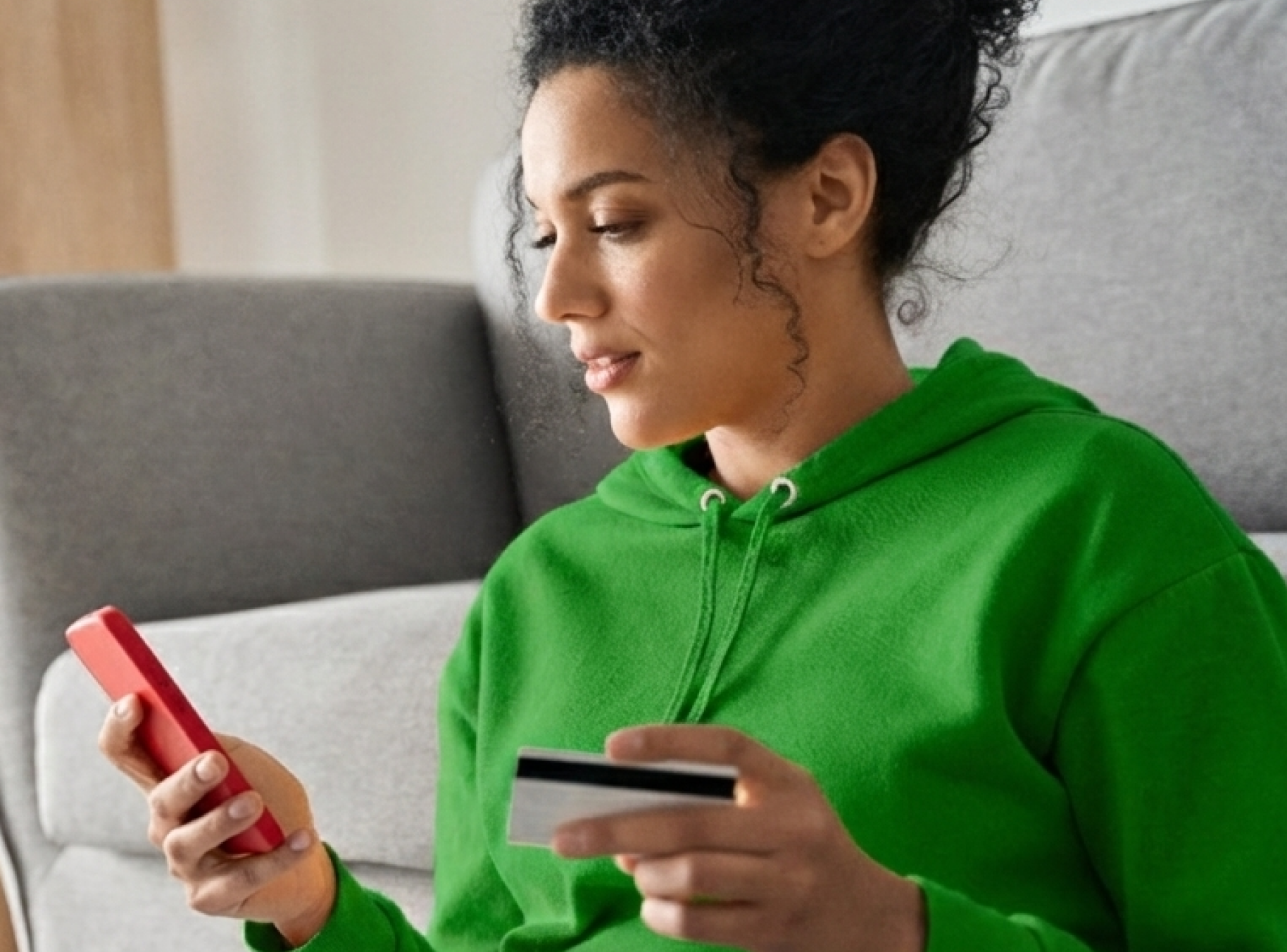 A woman in a green hoodie holds a smartphone and a credit card, sitting on a sofa at home. She appears focused, engaged in online shopping or banking.