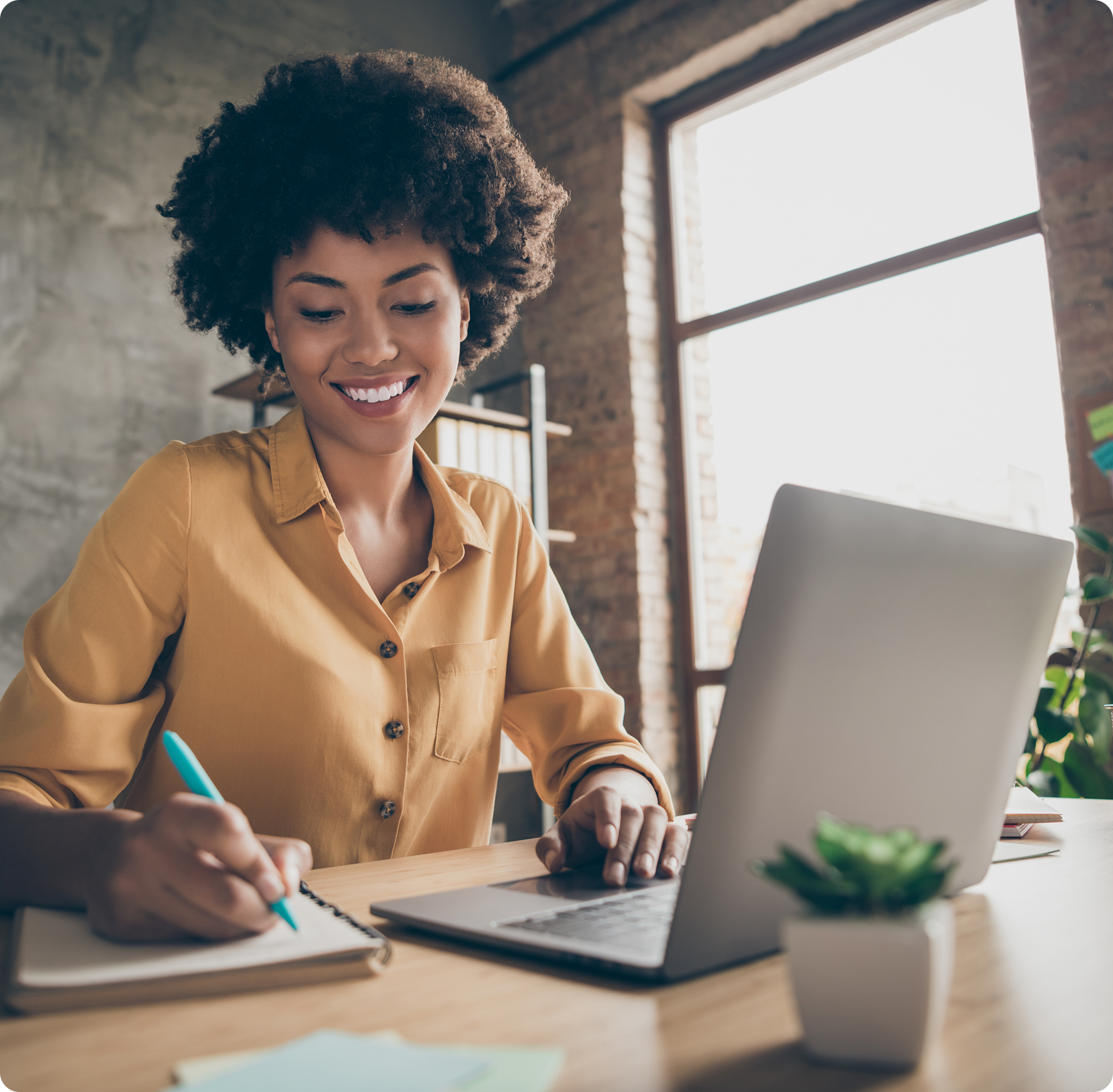 Smiling woman in a yellow shirt writes in a notebook beside an open laptop. She's working at a wooden desk near a large window, conveying a productive mood.