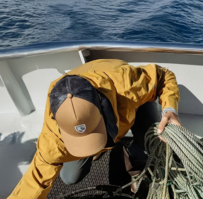 A person in a yellow jacket and cap organizes ropes on a boat deck against a backdrop of calm ocean and distant islands, conveying a sense of adventure.