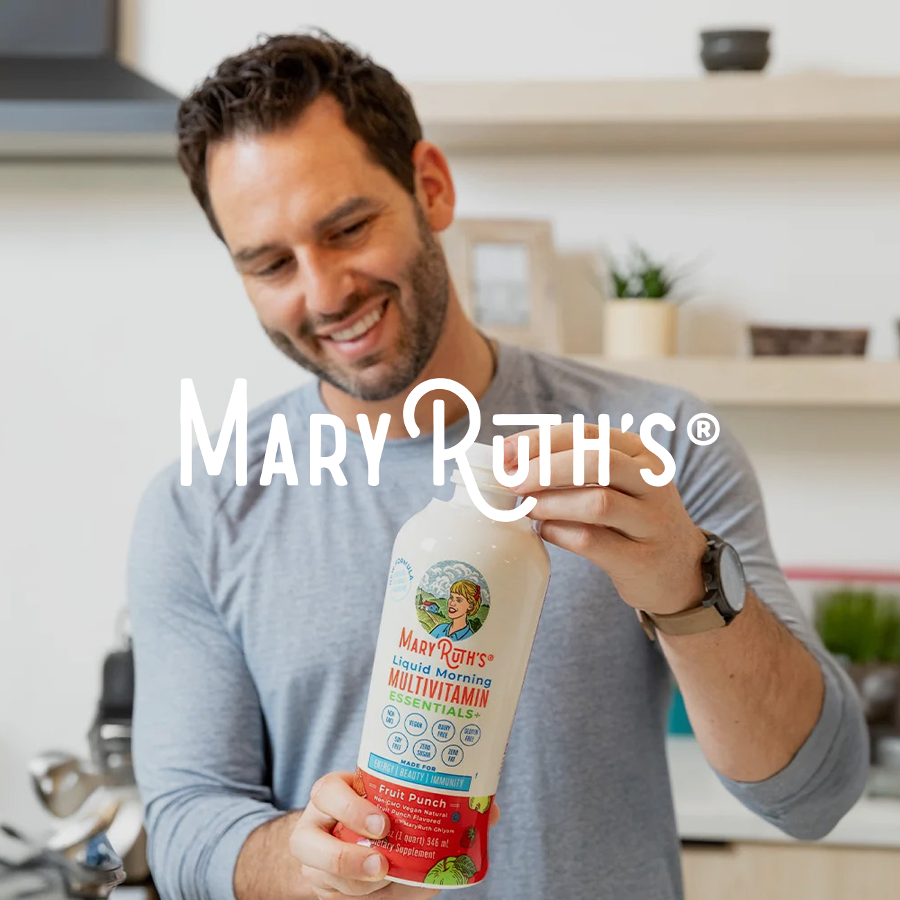 A smiling man holds a large bottle of Mary Ruth's Liquid Morning Multivitamin in a modern kitchen. The setting is bright and inviting, suggesting health and wellness.