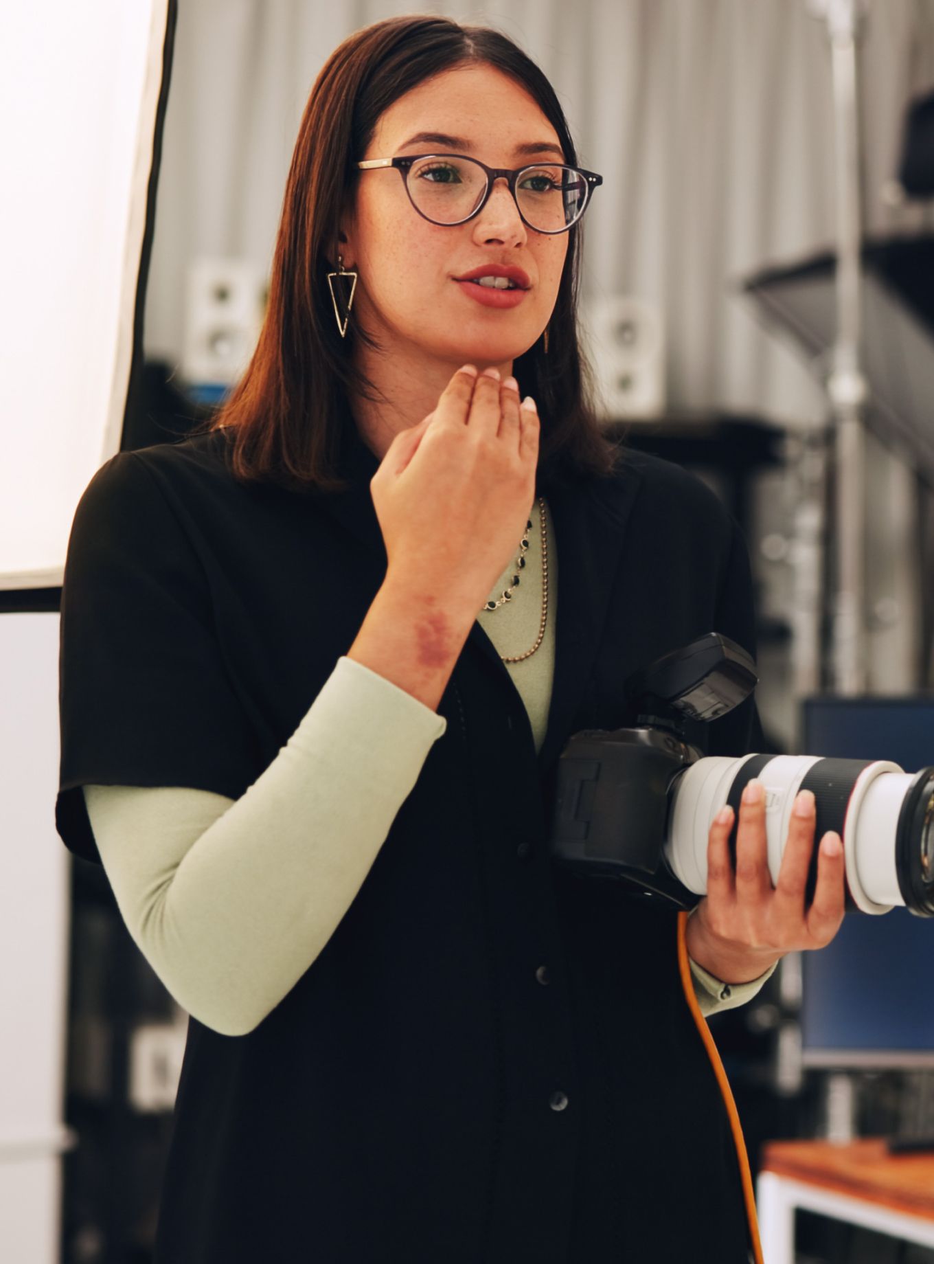 A woman with glasses and geometric earrings, wearing a black shirt over a light green top, holds a camera and gestures while speaking in a studio.