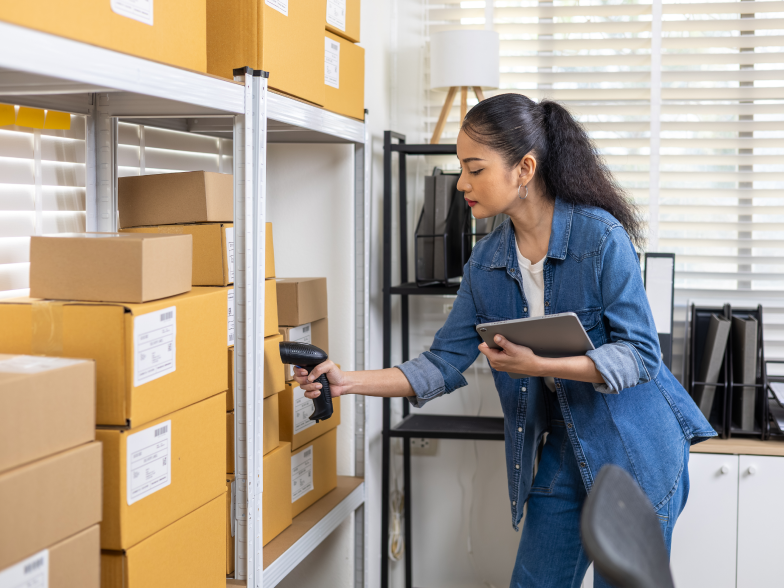 A small business owner scans a product with a handheld barcode reader while holding a tablet, standing at a shelving unit stocked with boxes near a sunlit window.
