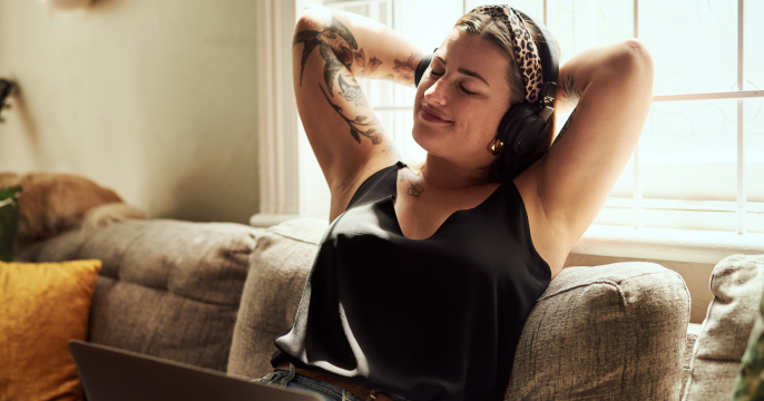 A person relaxes on a couch, wearing headphones and a leopard-print headband, eyes closed, smiling contentedly. Sunlight filters through a window. Tattoos on arms.