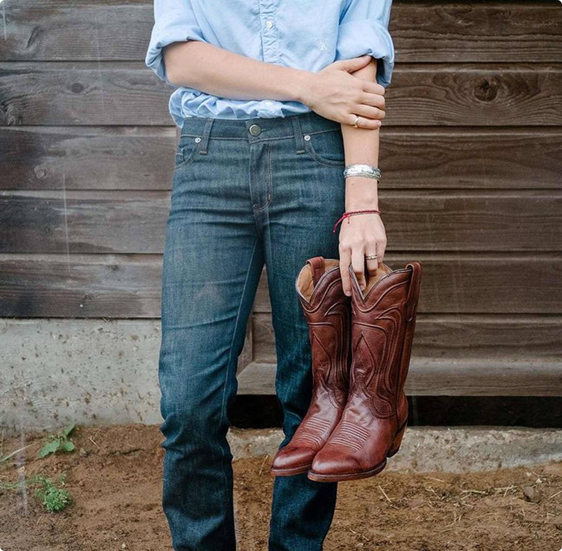 A person wearing a light blue shirt and jeans holds brown cowboy boots against a wooden wall. The scene has a rustic, casual feel.