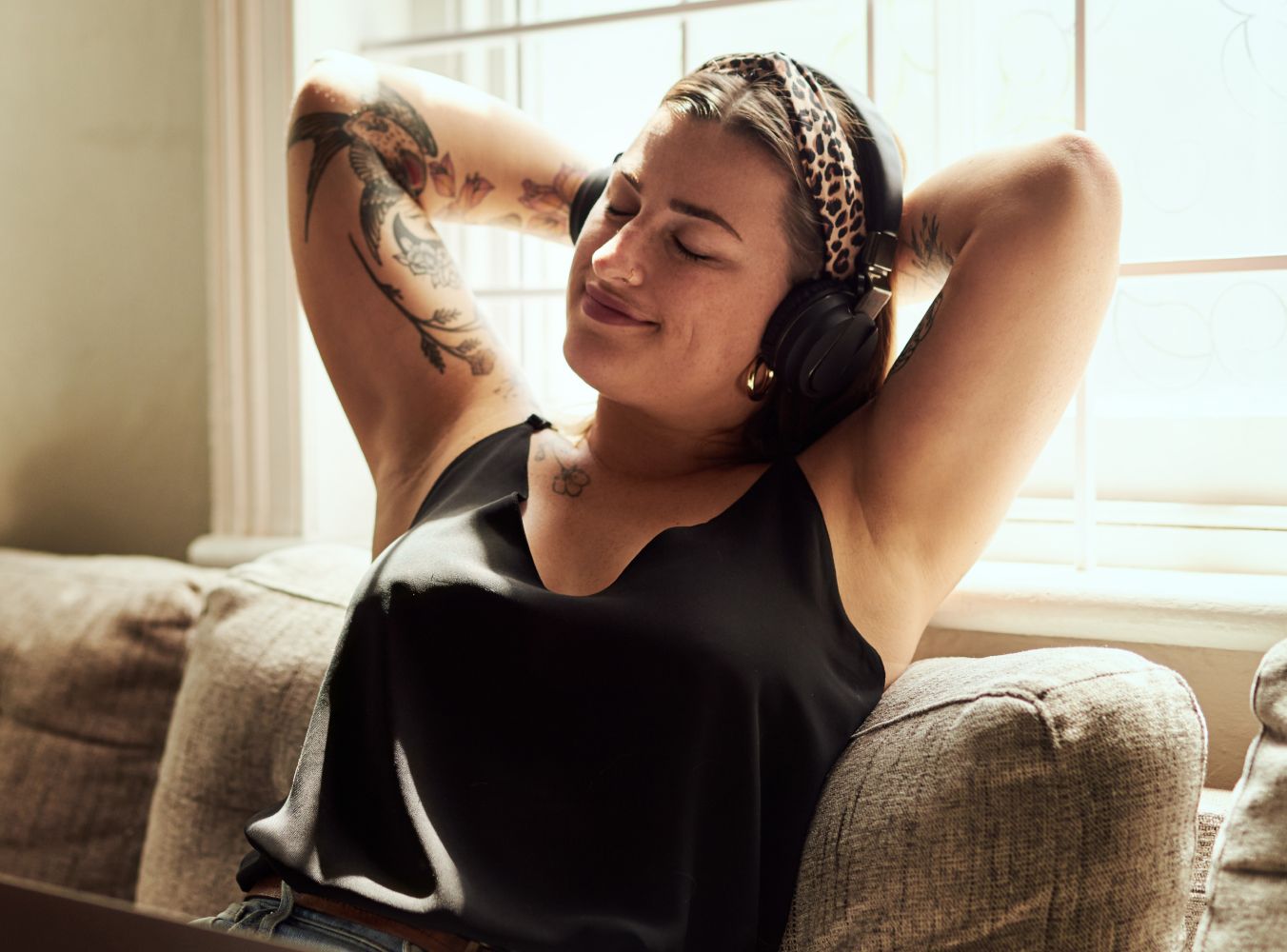 A person relaxes on a couch, wearing headphones and a leopard-print headband, eyes closed, smiling contentedly. Sunlight filters through a window. Tattoos on arms.
