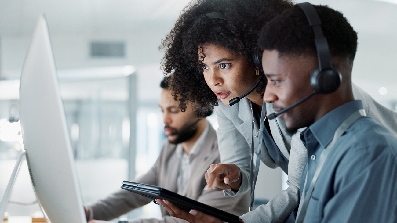 A focused woman in a headset points at a tablet, assisting a male colleague in an office. Another man works at a computer in the background.