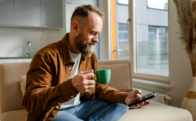 Man sits on a couch, smiling while looking at a smartphone and holding a green mug. The room is bright with natural light from a large window.