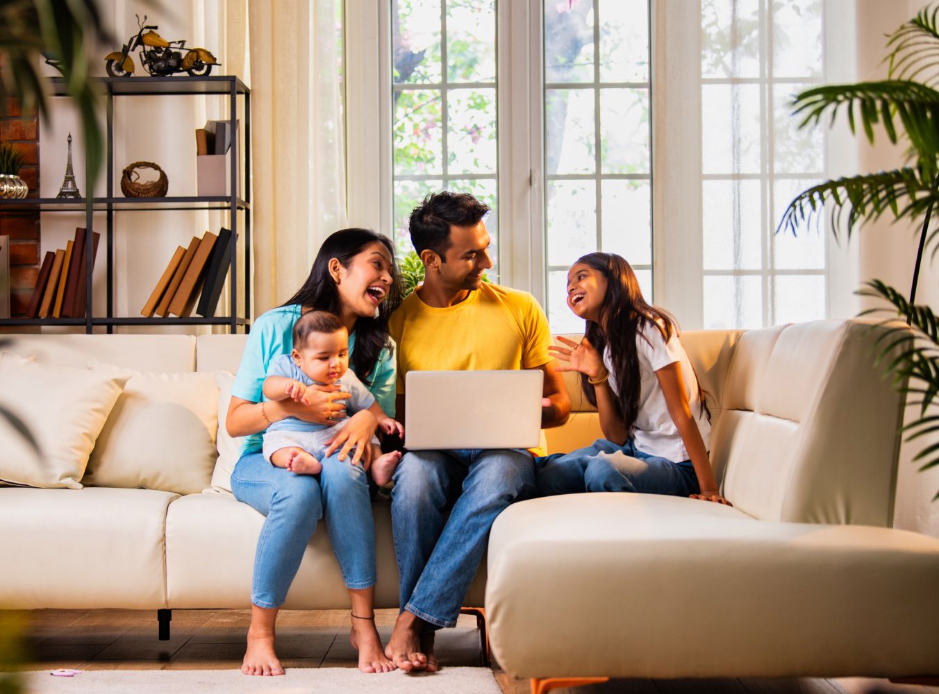 A joyful family of four sits on a beige sofa. Parents and two children gather around a laptop, smiling in a bright living room with large windows and plants.