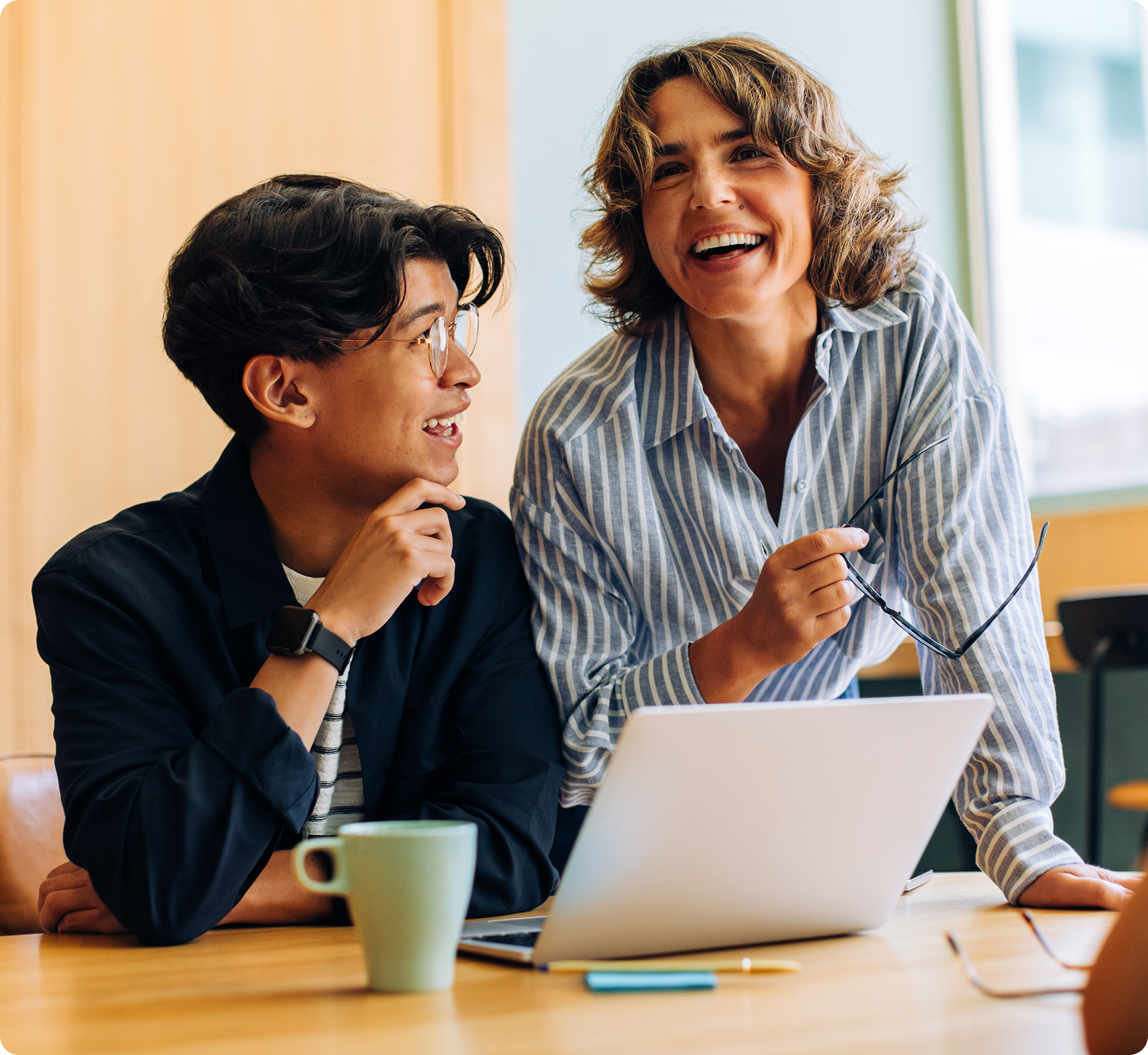 A cheerful woman and a young adult sit at a table with a laptop and coffee mug. They are laughing and appear to be having an engaging conversation.
