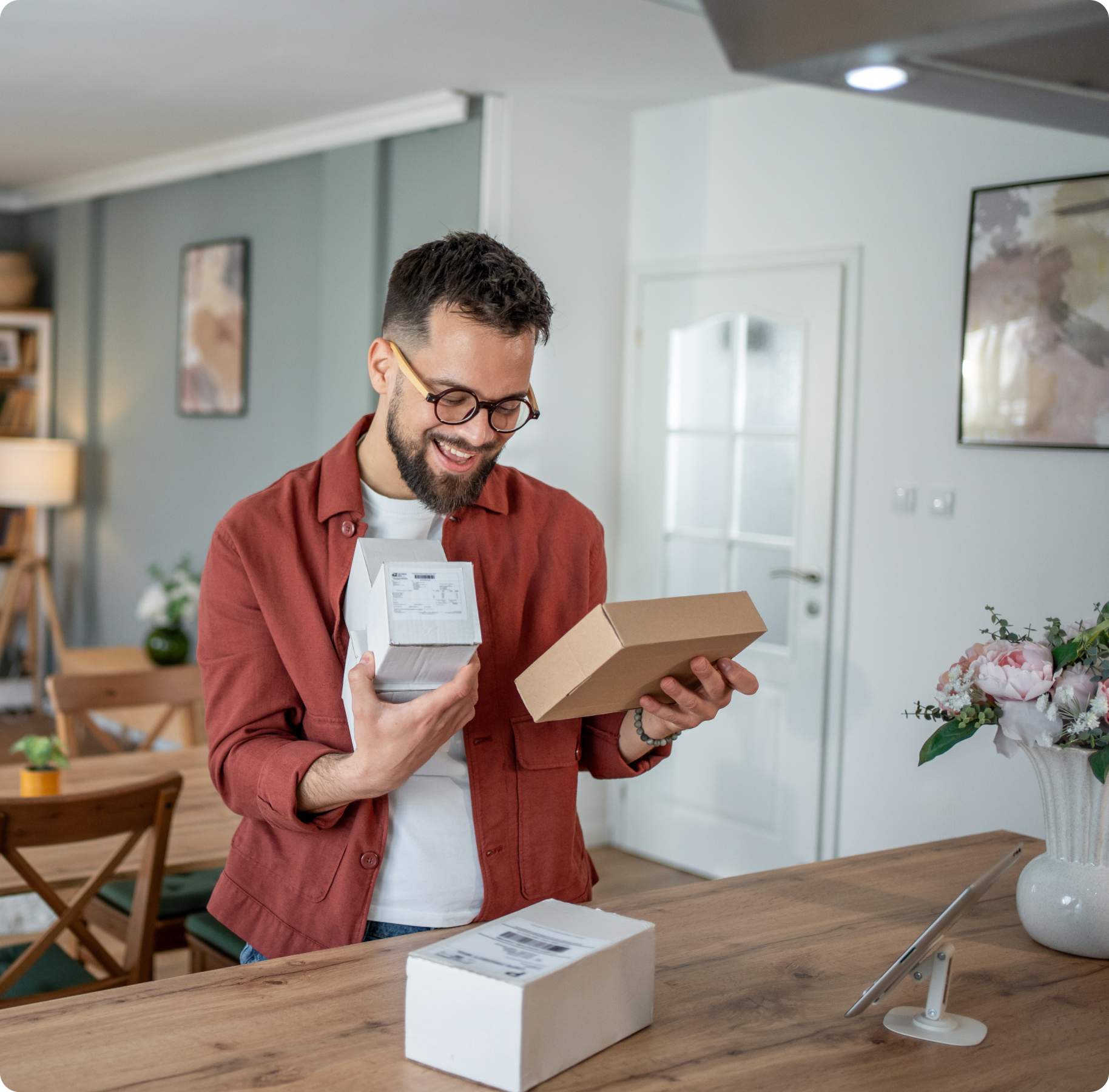 A man with glasses smiles while holding a small package in a cozy, well-lit living room. The room features wooden furniture and decorative plants, creating a warm atmosphere.