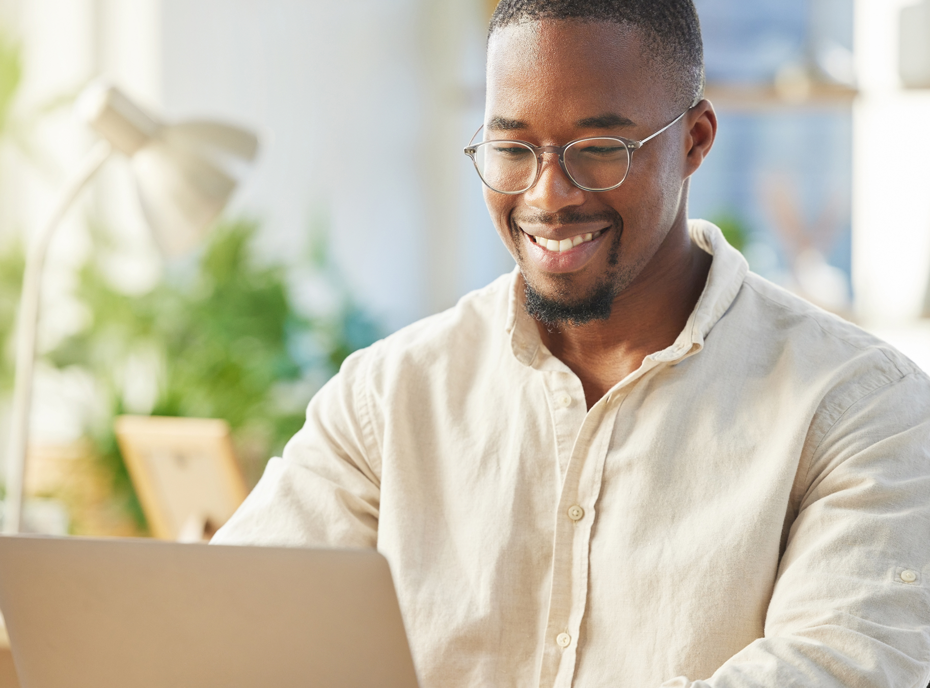 Smiling man with glasses using a laptop in a bright, plant-filled office. The scene conveys a positive and productive work atmosphere.