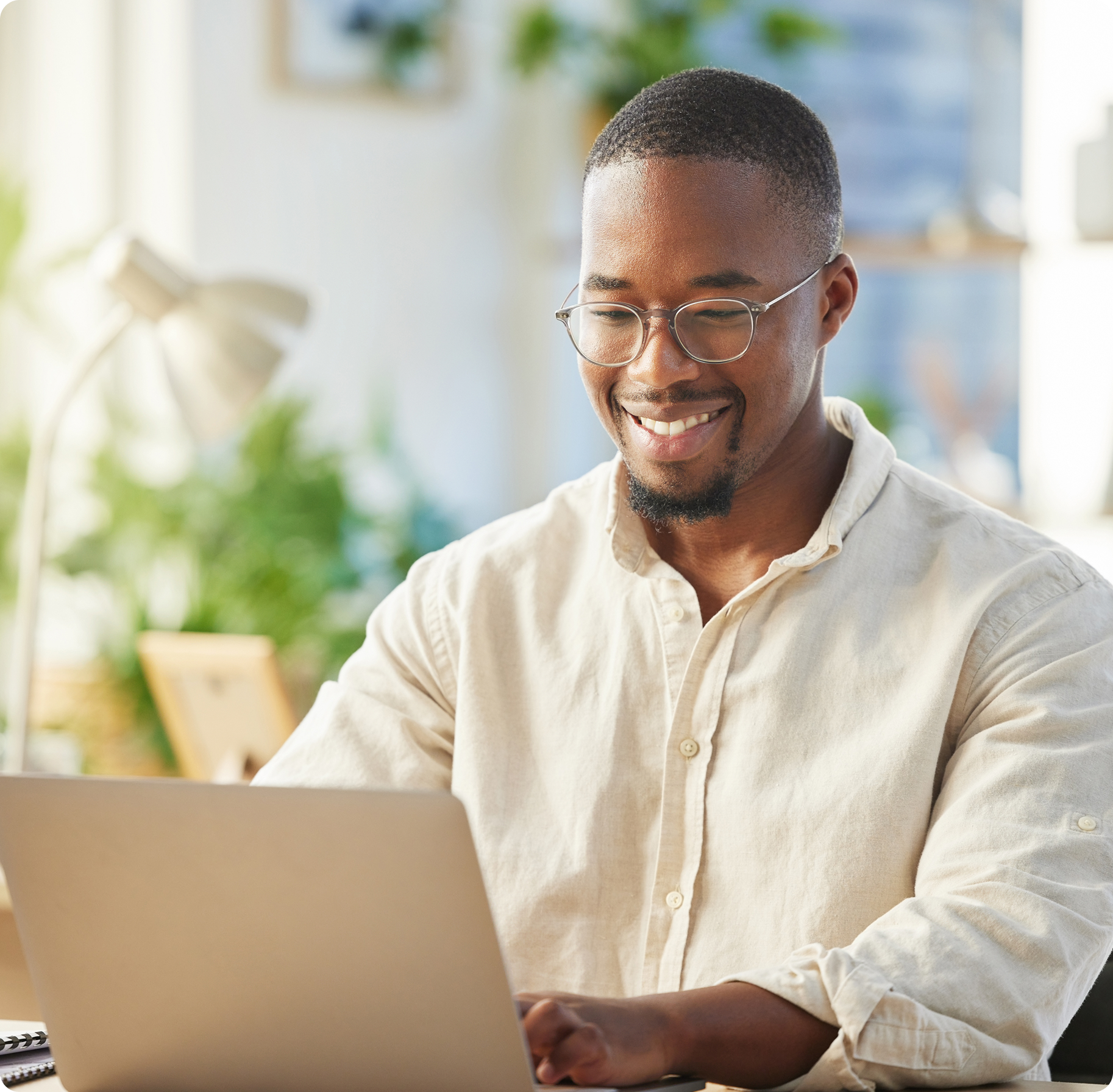 Smiling man with glasses using a laptop in a bright, plant-filled office. The scene conveys a positive and productive work atmosphere.