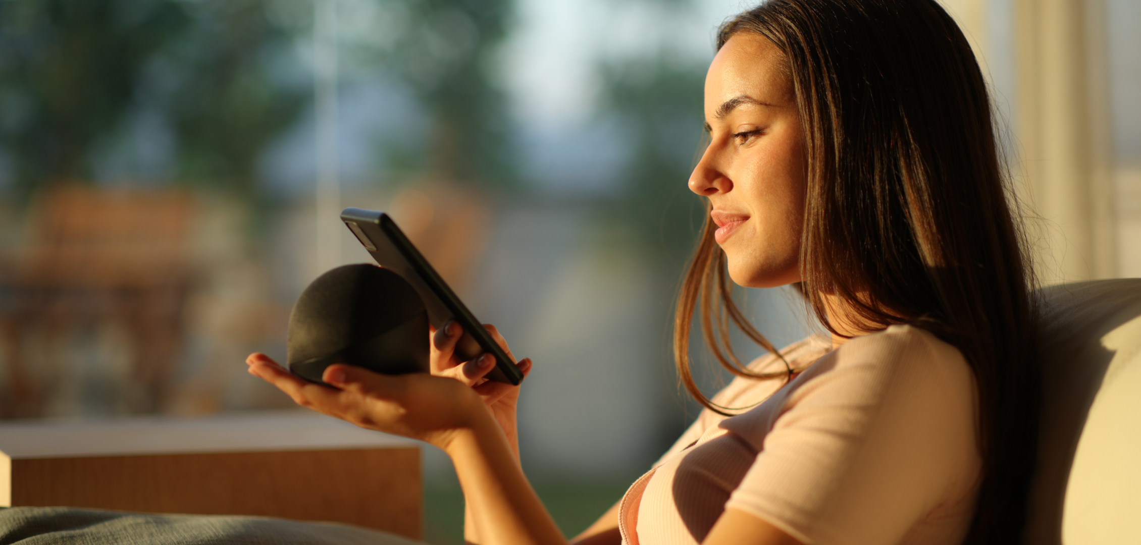 A young woman sits on a couch, holding a smartphone and a smart speaker. She looks relaxed and content, with sunlight gently illuminating her face.