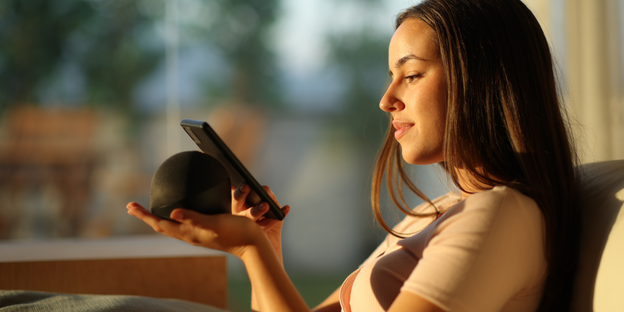 A young woman sits on a couch, holding a smartphone and a smart speaker. She looks relaxed and content, with sunlight gently illuminating her face.
