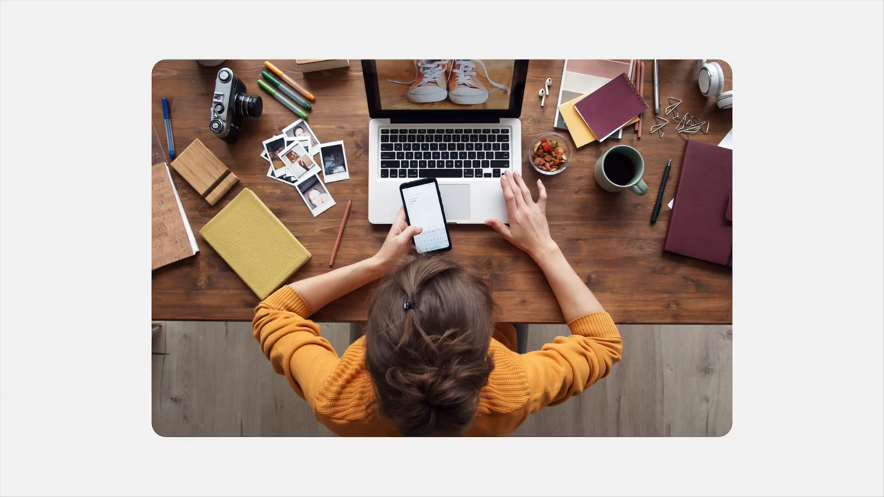 A person in a yellow sweater sits at a wooden desk with a laptop, smartphone, notebooks, camera, and coffee, creating a busy, creative workspace.