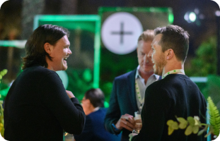 Three men are engaged in a lively conversation at an outdoor event, surrounded by greenery and soft evening lighting, creating a warm and friendly atmosphere.