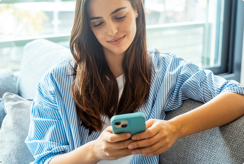 A woman in a blue striped shirt sits on a couch near a window, smiling as she looks at her smartphone. The scene is relaxed and casual, conveying a sense of contentment.
