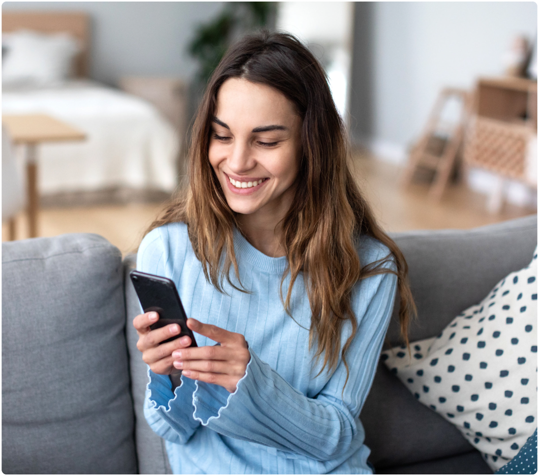 A smiling woman in a blue sweater sits on a couch using a smartphone in a cozy, well-lit room. The atmosphere is relaxed and cheerful.