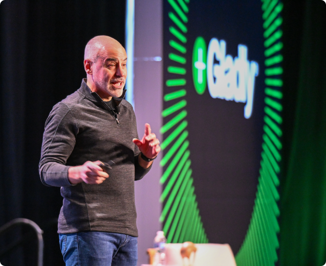 A man speaks energetically on stage, wearing a gray sweater and jeans. A large screen behind him displays the word "Gladly" with a dynamic green design.
