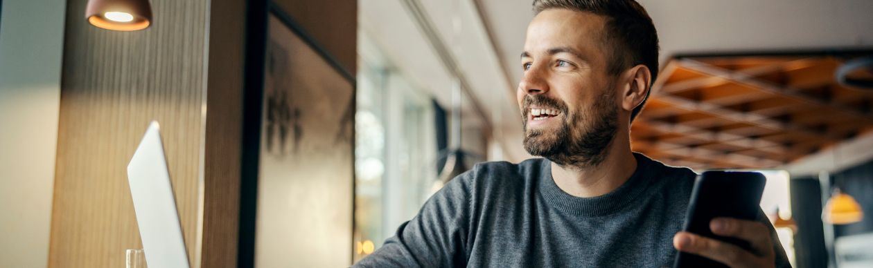 Bearded man smiling and holding a smartphone in front of a laptop