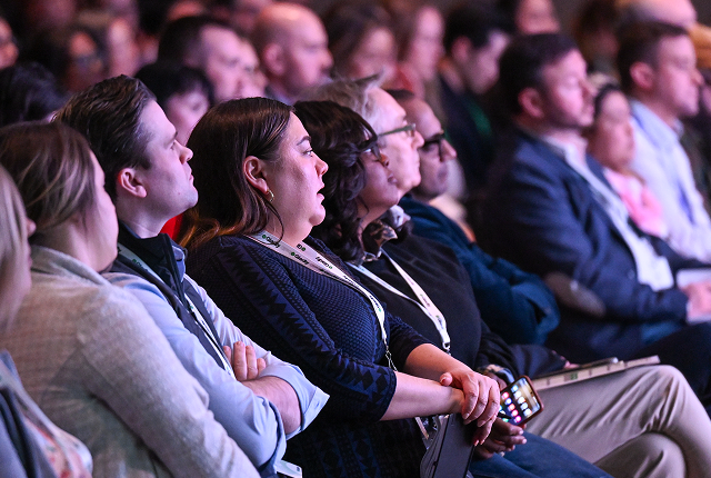 Audience attentively watching a presentation, seated in rows. Mixed emotions of focus and engagement are visible. The lighting is soft and ambient.