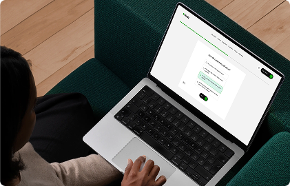 A person sits on a dark green couch using a laptop displaying a survey form. The wooden floor and relaxed posture create a focused yet casual atmosphere.