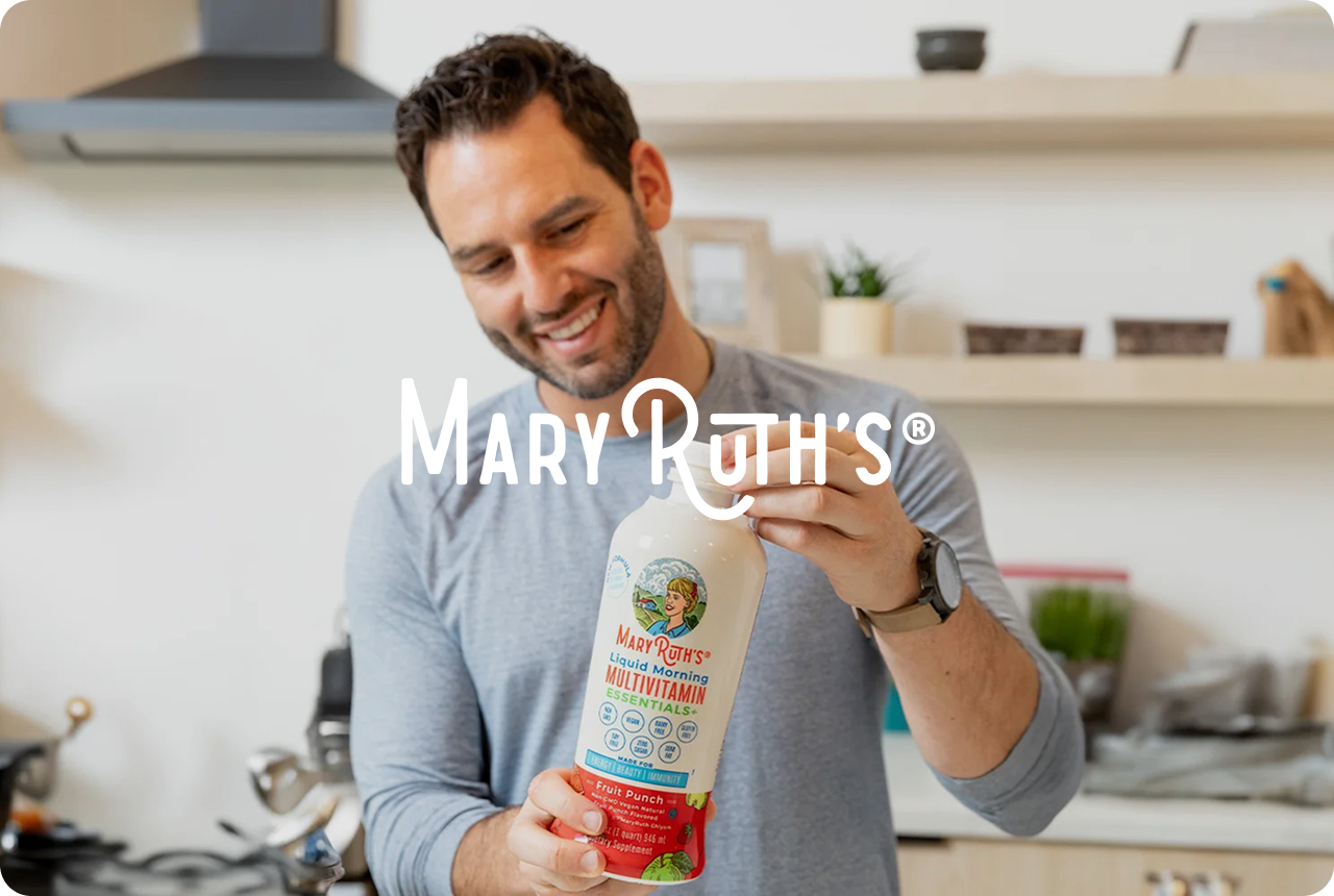 A smiling man holds a MaryRuth's multivitamin bottle in a modern kitchen, conveying health and wellness. Shelving and kitchenware are in the background.