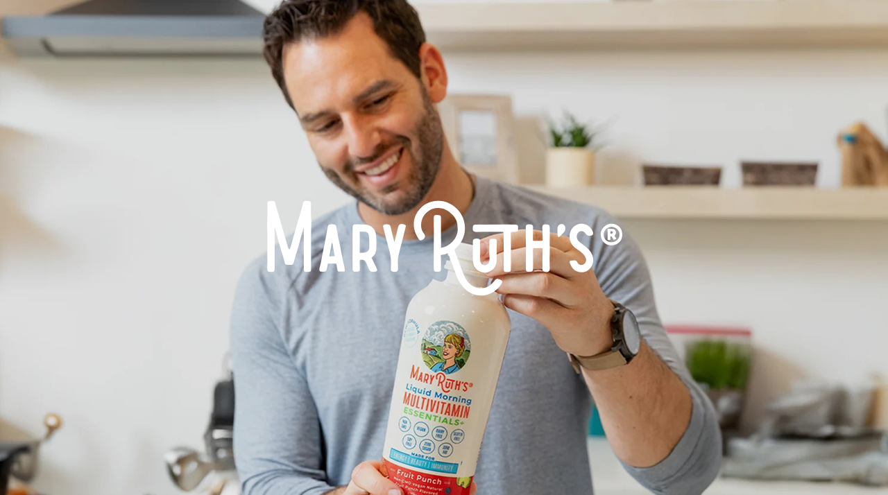 A smiling man holds a MaryRuth's multivitamin bottle in a modern kitchen, conveying health and wellness. Shelving and kitchenware are in the background.