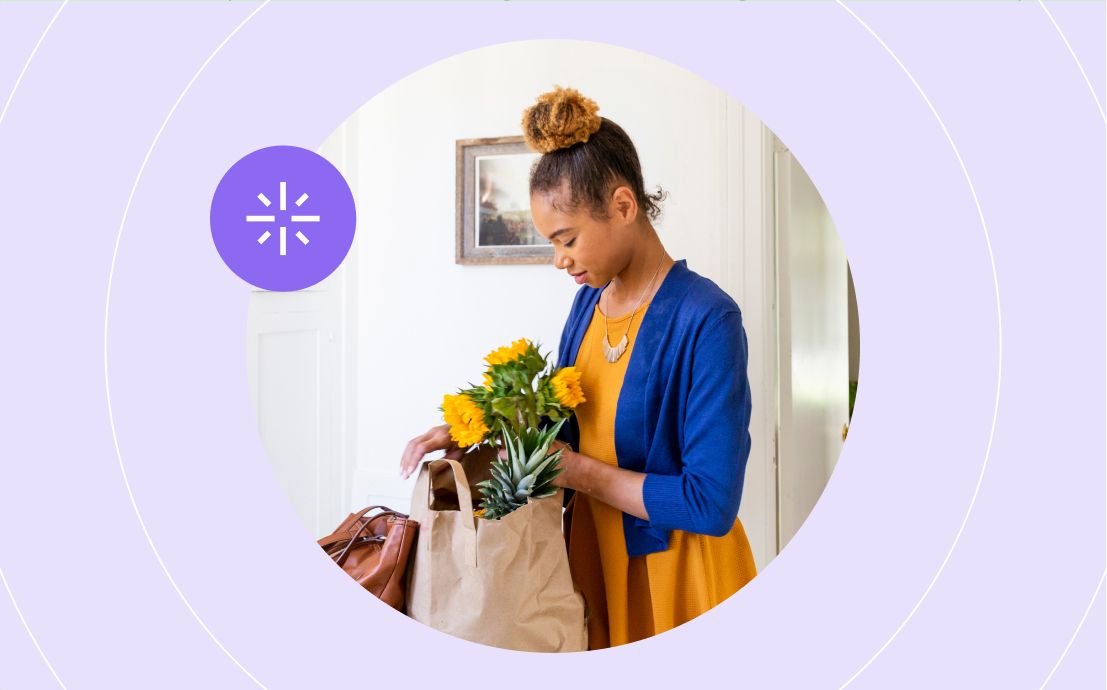 A woman in a blue cardigan and yellow dress places sunflowers and produce into a paper bag at home. The mood is calm and organized.
