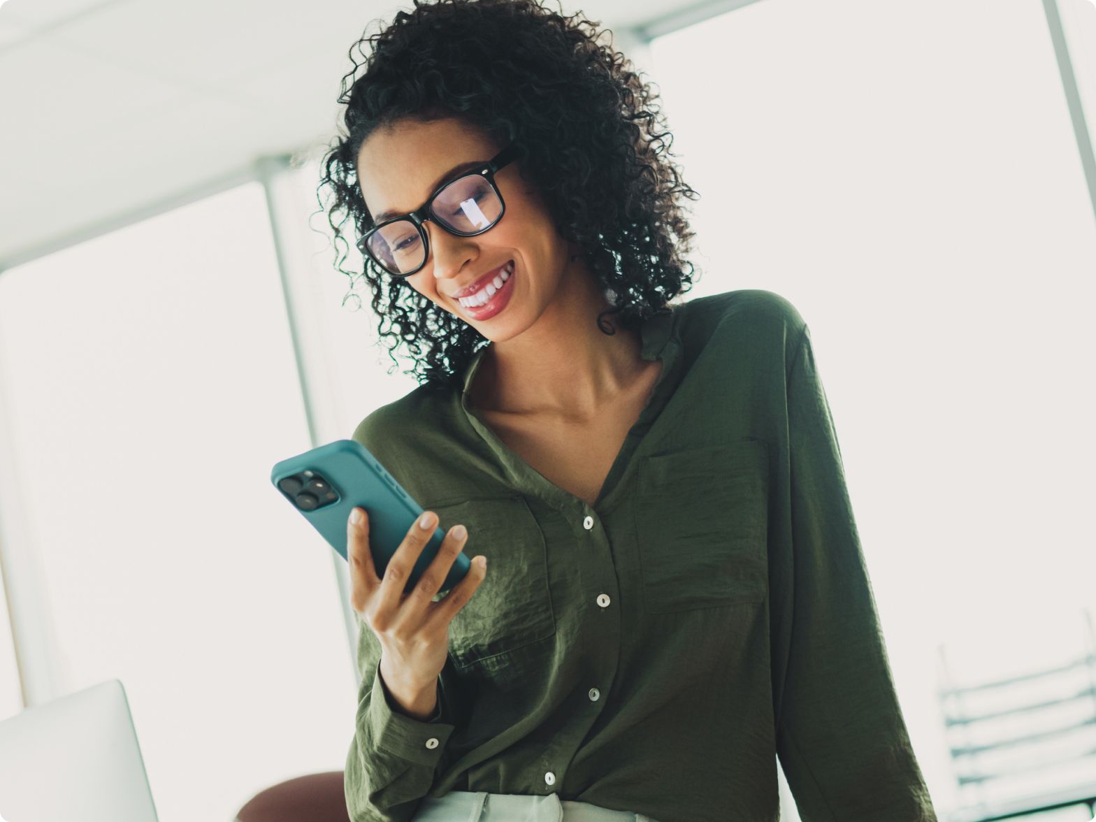 A woman with curly hair and glasses smiles while looking at her smartphone. She is wearing a green blouse, and the background is a bright office space.