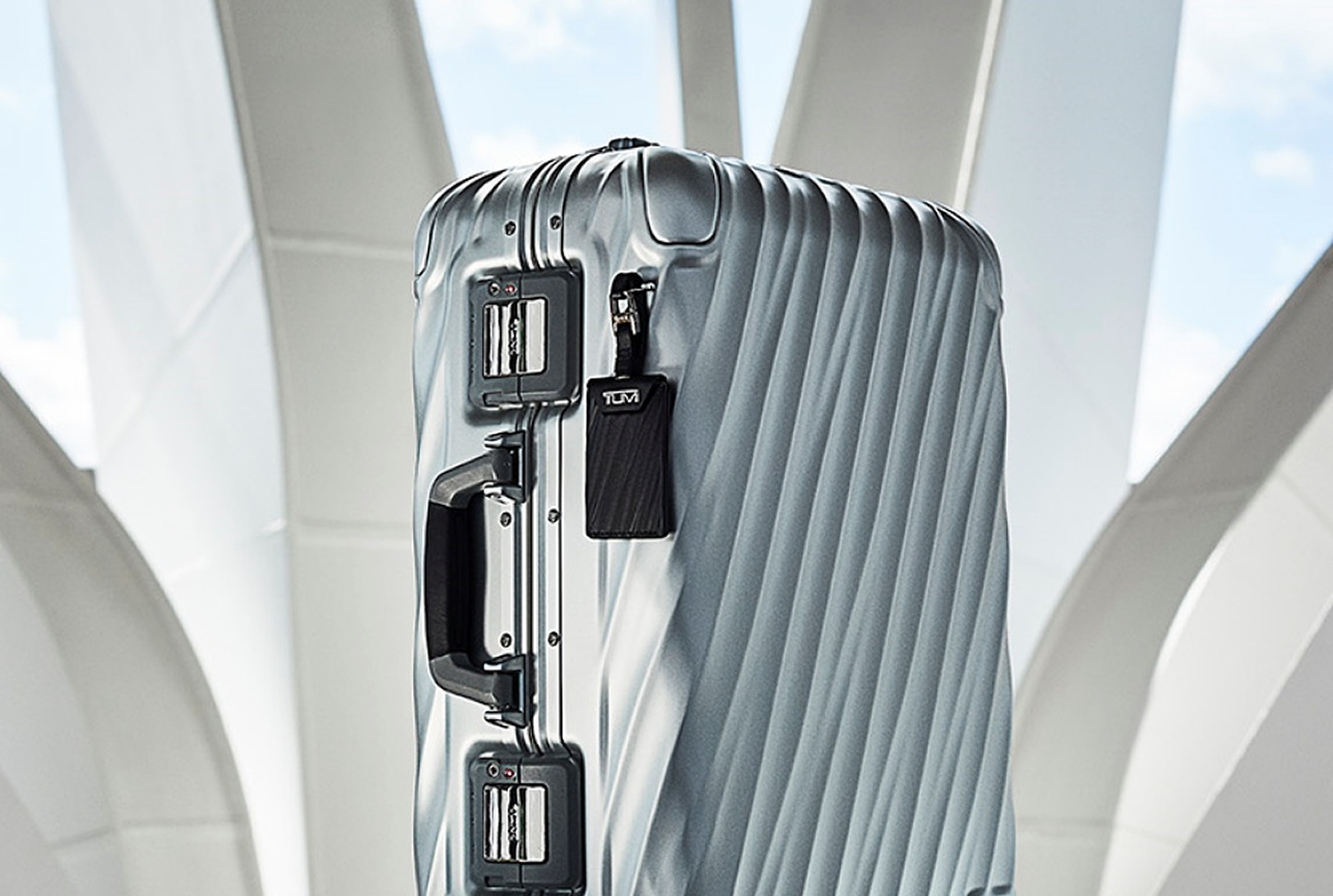 A sleek silver suitcase with a textured surface and black handles stands on four wheels. It’s set against a modern architecture backdrop, suggesting travel.