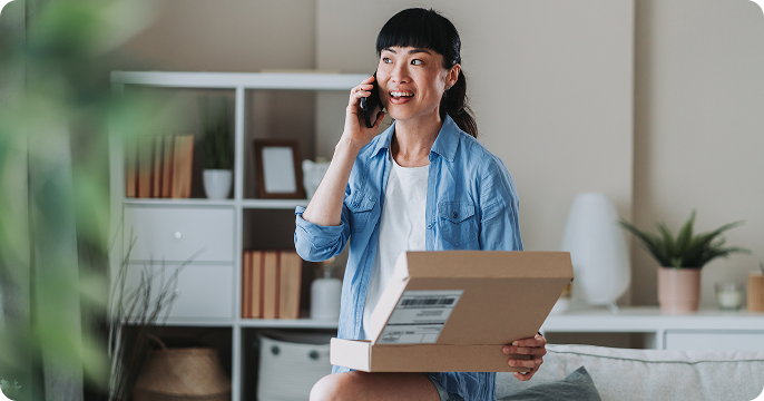 A woman smiles while talking on the phone, holding an open cardboard box in a cozy living room. She wears a blue shirt and white top, with shelves in the background.