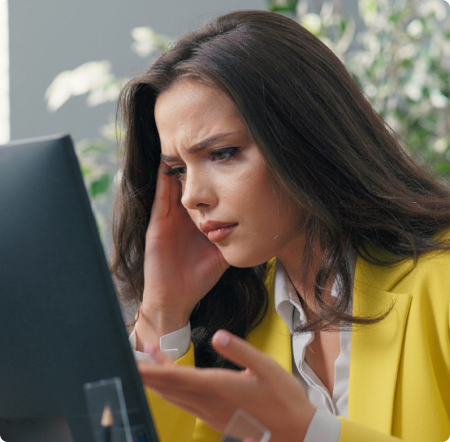 A woman in a yellow blazer looks frustrated while staring at a computer screen, resting her head on her hand. Plants are blurred in the background.