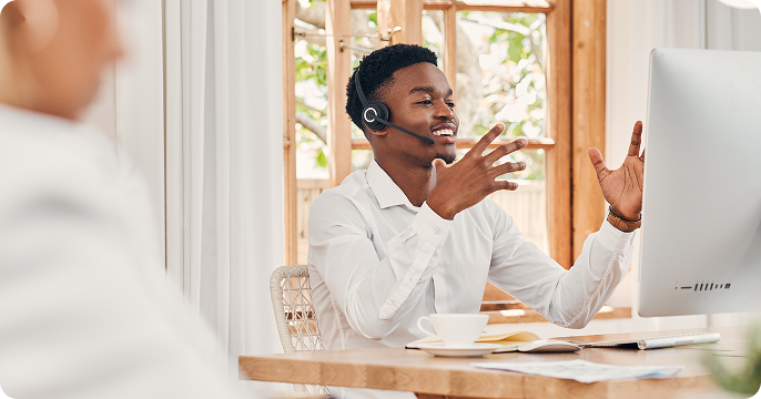 Young professional wearing a headset, smiling and gesturing while on a video call at a desk with a computer and coffee. Bright, airy office setting.