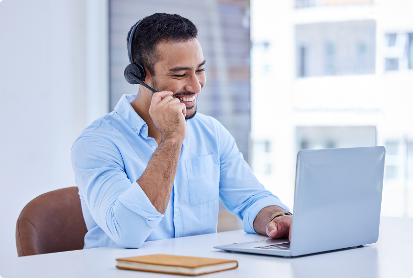 Smiling man in a light blue shirt with a headset, engaged in a call, sitting at a desk with a laptop. Bright, professional setting conveys positivity.