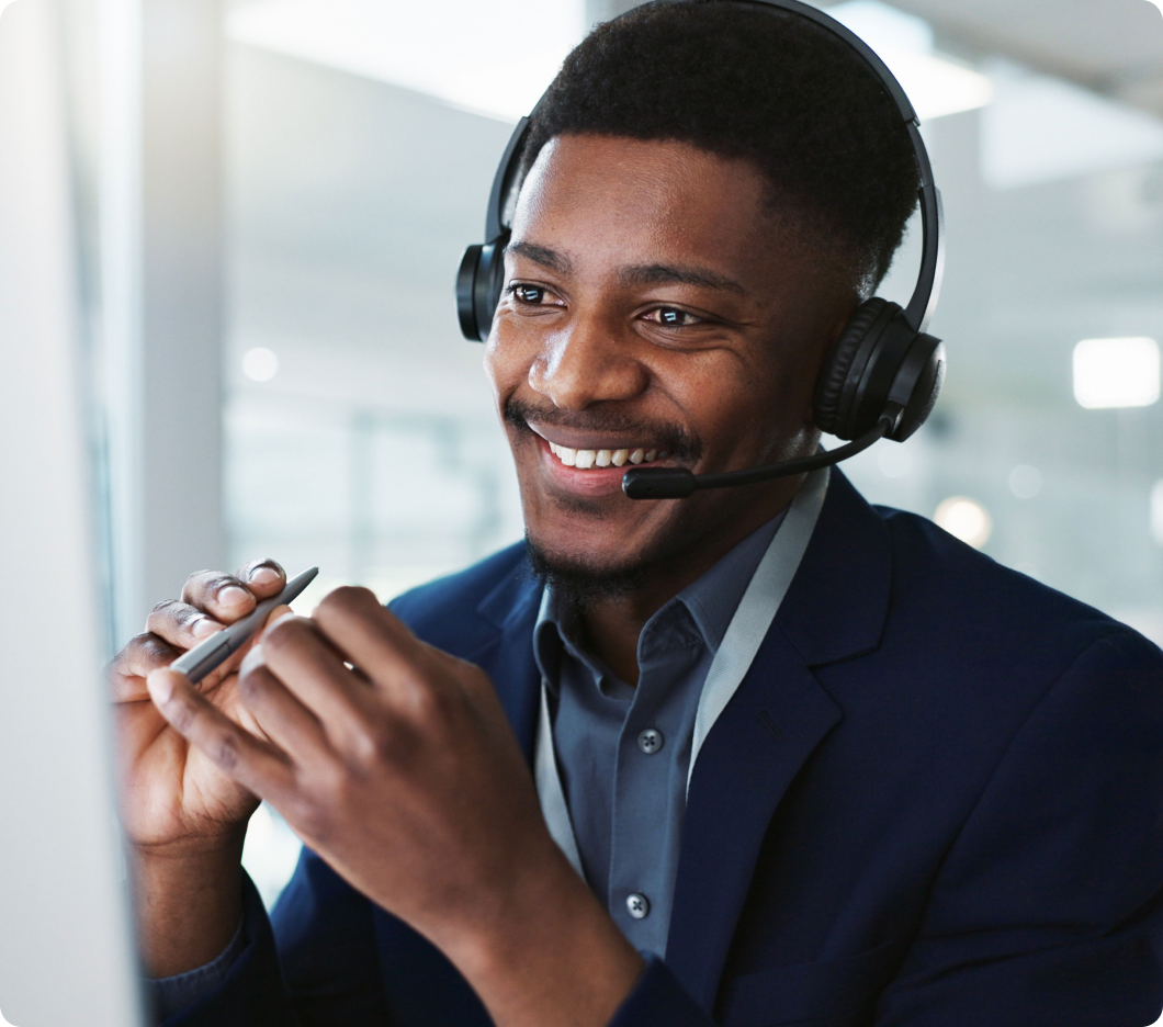 Smiling man with a headset, wearing a suit in a bright office, gestures with a pen during a video call, conveying professionalism and friendliness.