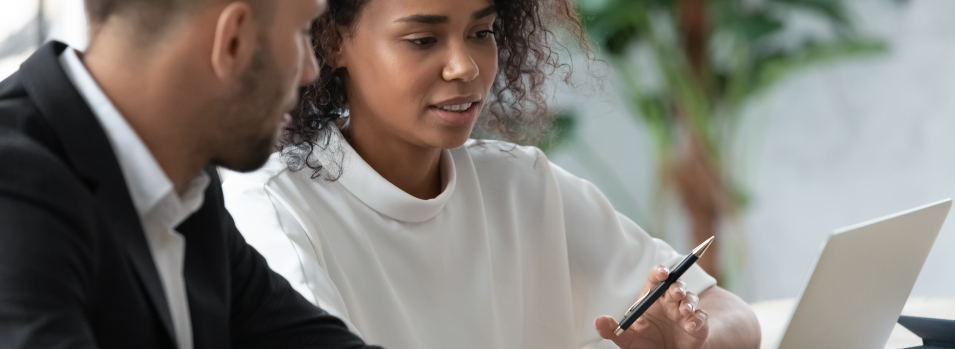 A customer success manager gestures with a pen toward a laptop screen as she walks a client through an account review at a table in a bright office