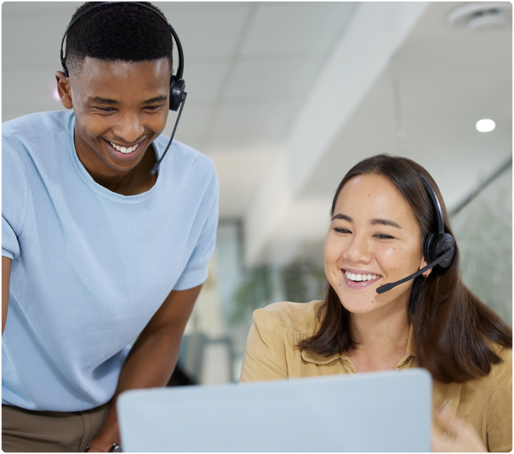 Two people, both wearing headsets, are smiling and looking at a laptop screen. The setting appears to be a modern office, conveying a positive work environment.