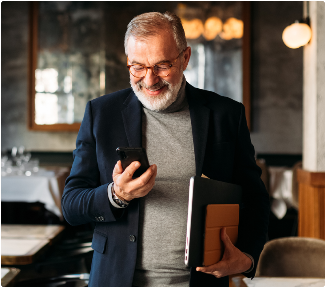 Elderly man with glasses smiling at phone in restaurant, holding a laptop. He wears a dark blazer and gray turtleneck. Warm, inviting atmosphere.