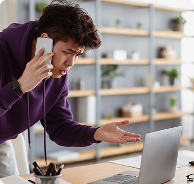 A man in a purple hoodie, looking frustrated, holds a smartphone to his ear while gesturing at a laptop. The setting is a modern home with shelves.