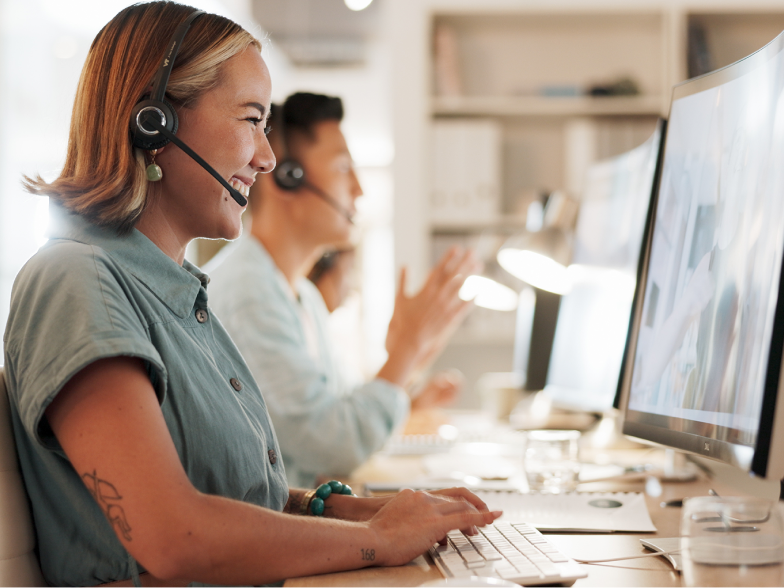 A smiling live chat agent wearing a headset types a response to a customer on her keyboard in a bright, modern support office, with a teammate helping customers in the background
