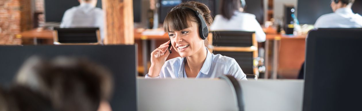 A woman in bangs wearing a phone headset sitting in a customer service office and smiling