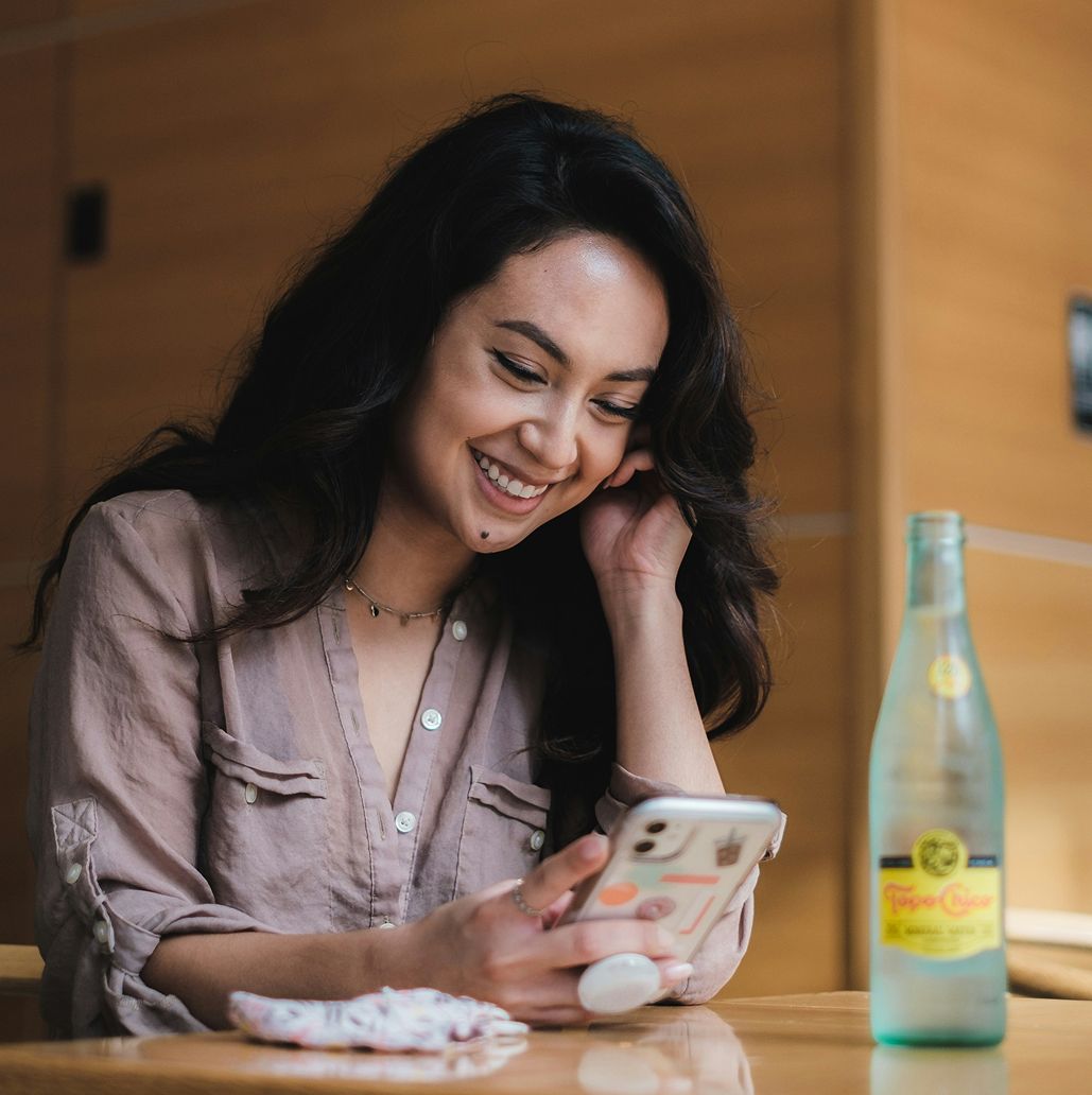 A woman in a cafe smiles while texting on her phone. A chat bubble reads, “Hey! I love the jacket, but I’m thinking I should've gone with the black instead. Can I swap it?” Another bubble replies, “Absolutely—let me kick off the exchange for black in your size.” The scene conveys a friendly, relaxed mood.